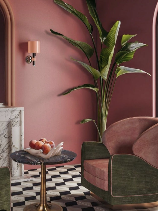 Elegant interior with a pink wall, green velvet chair, marble side table holding a bowl of peaches, and a large potted plant, set against a checkered floor.