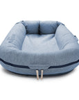 A square-shaped, denim-blue dog bed with raised padded edges and a darker blue trim. The bed features a subtle centered DockATot logo and conforms to the Oeko-Tex Standard 100, ensuring it is LAST CHANCE: Deluxe+ Dock - Indigo Chambray.