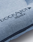 Close-up of a blue DockATot Deluxe+ Dock - Indigo Chambray multifunctional docking station showing the brand name embossed in gray on the fabric, with a focus on the textured material and the visible zipper detail along its edge.