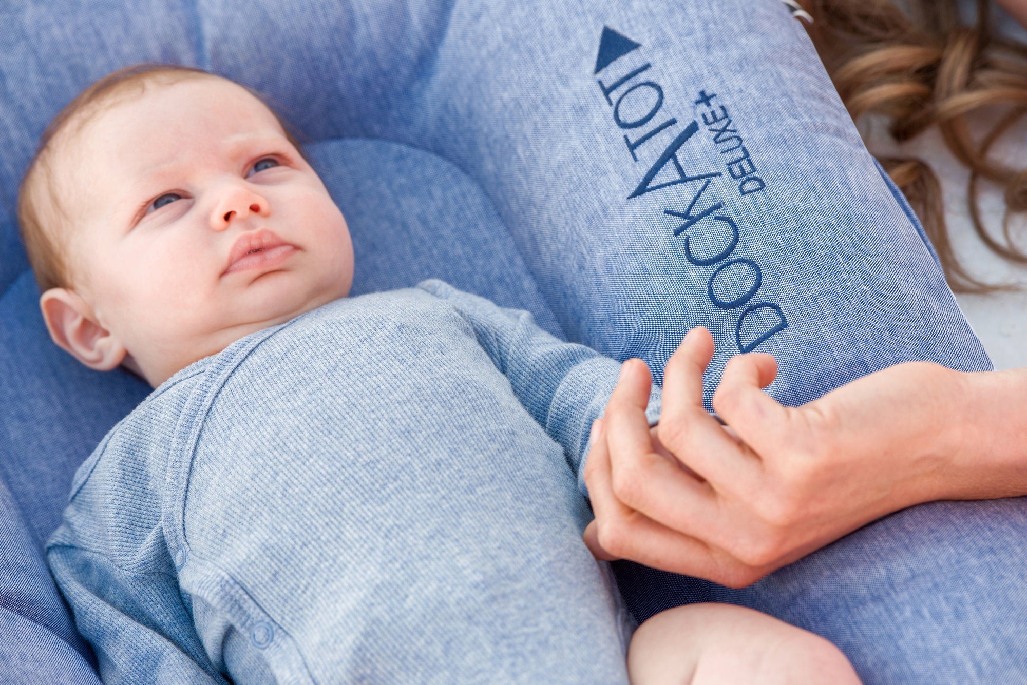 A newborn baby dressed in a blue outfit lies on a gray, cushioned DockATot Deluxe+ Dock - Indigo Chambray, held gently by a woman&#39;s hands, with partial views of her flowing brown.