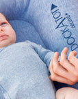 A newborn baby dressed in a blue outfit lies on a gray, cushioned DockATot Deluxe+ Dock - Indigo Chambray, held gently by a woman's hands, with partial views of her flowing brown.