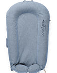 A blue DockATot LAST CHANCE: Deluxe+ Dock - Indigo Chambray baby lounger isolated on a white background. The lounger has a cushioned, oval shape with side handles and prominently displays the "DockATot" logo.