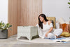 A woman sits on a round beige rug in a bright room, looking at an open book next to a DockATot Kind Essential Bassinet - Willow Boughs with ornately crafted pedestal feet. Wooden panels and a plant.